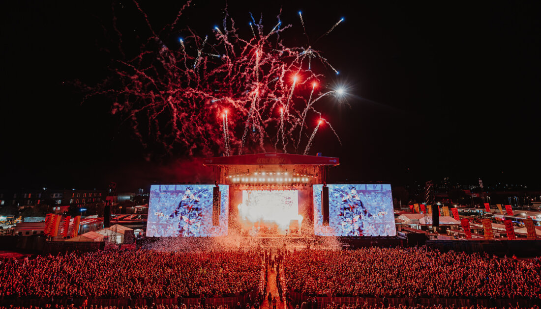 A vibrant music festival scene at night featuring a large crowd, fireworks, and the main stage illuminated with colorful lights.