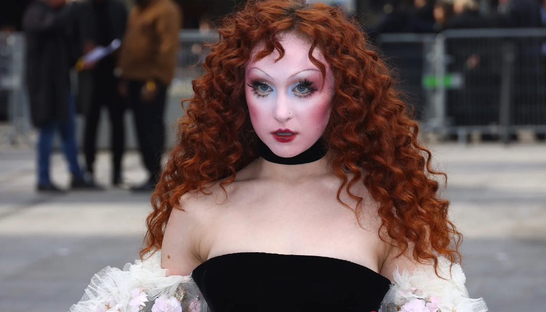 Chappell Roan wears a cream coloured lace skirt on top of a black corset top and red bow tied around her waist. She has bright red auburn hair styled in waves 