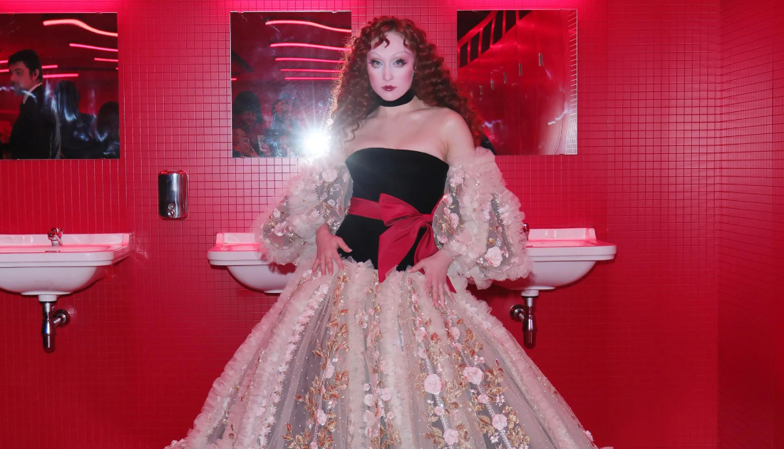 Chappell Roan wears a cream coloured lace skirt on top of a black corset top and red bow tied around her waist. She has bright red auburn hair styled in waves 