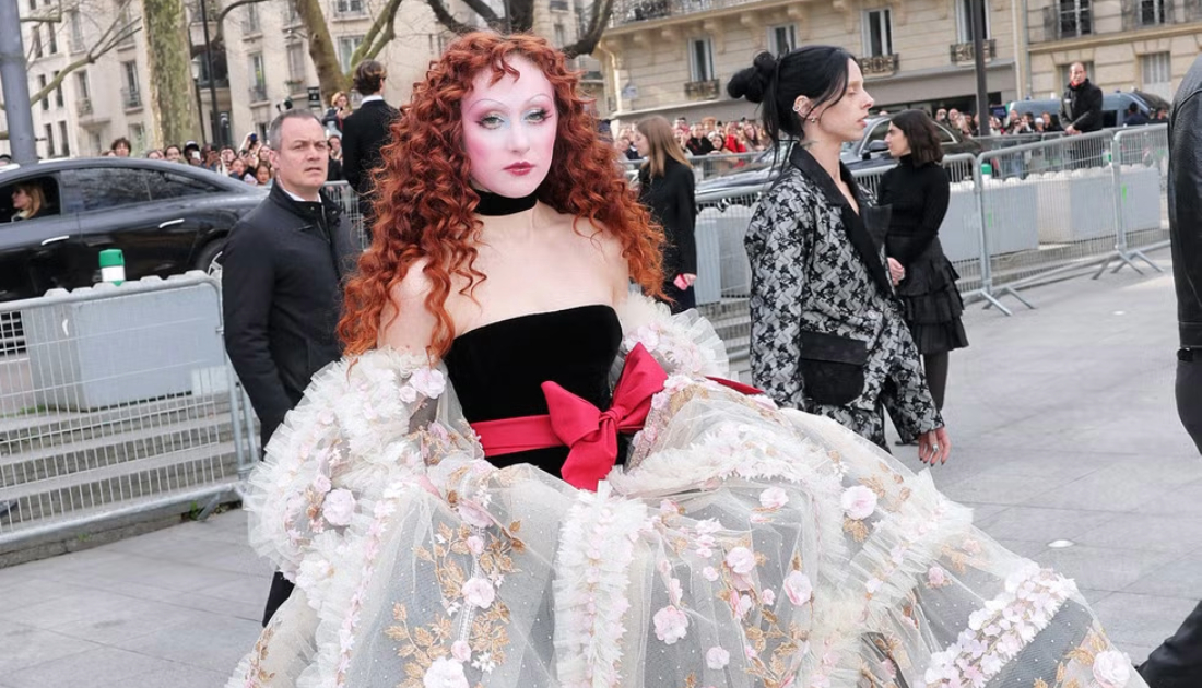 Chappell Roan wears a cream coloured lace skirt on top of a black corset top and red bow tied around her waist. She has bright red auburn hair styled in waves 
