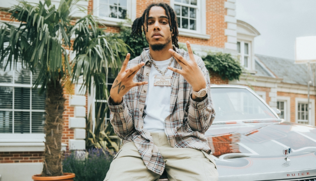 Artist AJ Tracey sitting on top of car parked outside a mansion 